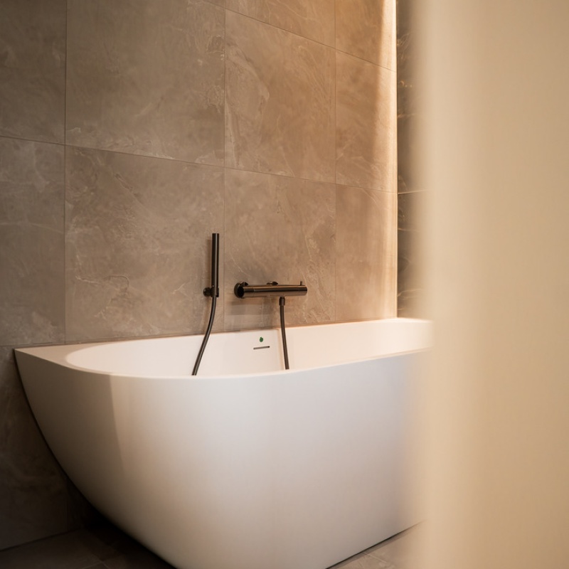 Freestanding white bathtub with bronze wall-mounted faucet in the renovated bathroom of the Geuzenkwartier home in The Hague, finished with large grey-beige tiles and indirect lighting